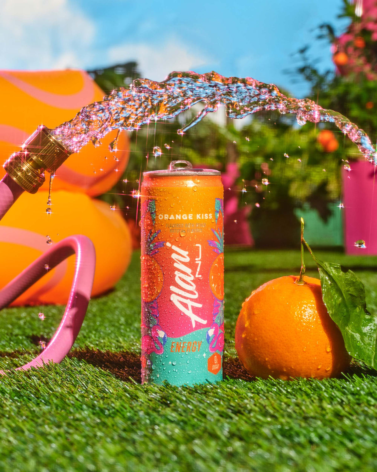A pink garden hose pouring water over a citrus fruit and an Orange Kiss Energy Drink.
