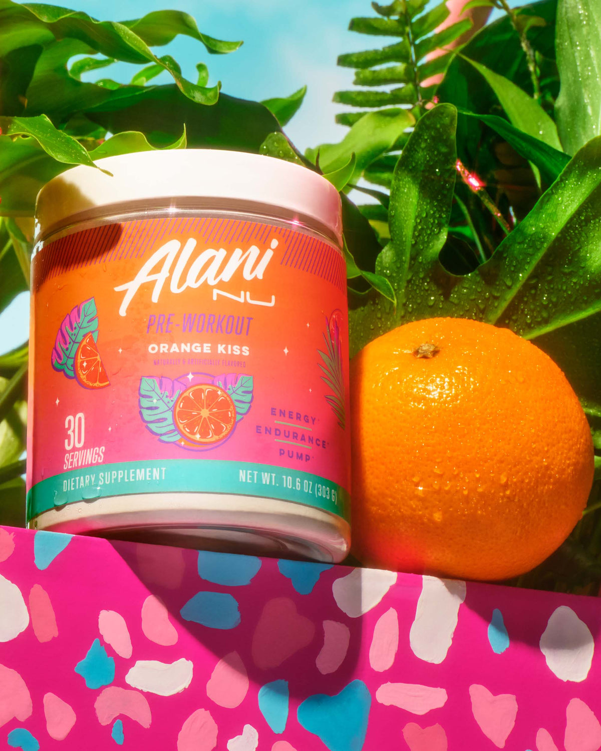 An orange and a tub of Pre-Workout in front of dewy tropical leaves, atop a pink terrazzo counter.