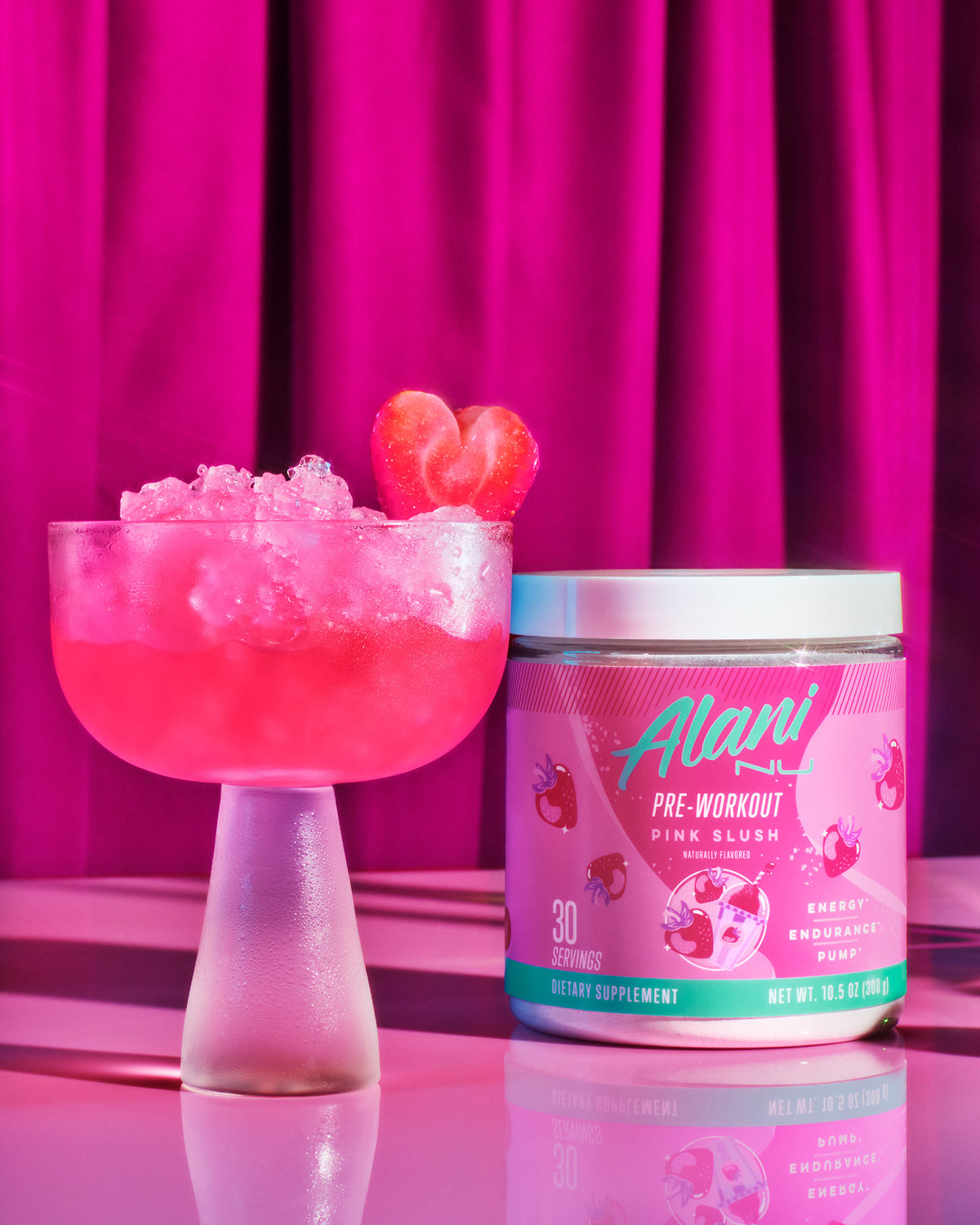 A tub of Alani Nu Pink Slush Pre-Workout next to a frosted coup glass filled with pebble ice and pink liquid garnished with a heart-shaped strawberry.