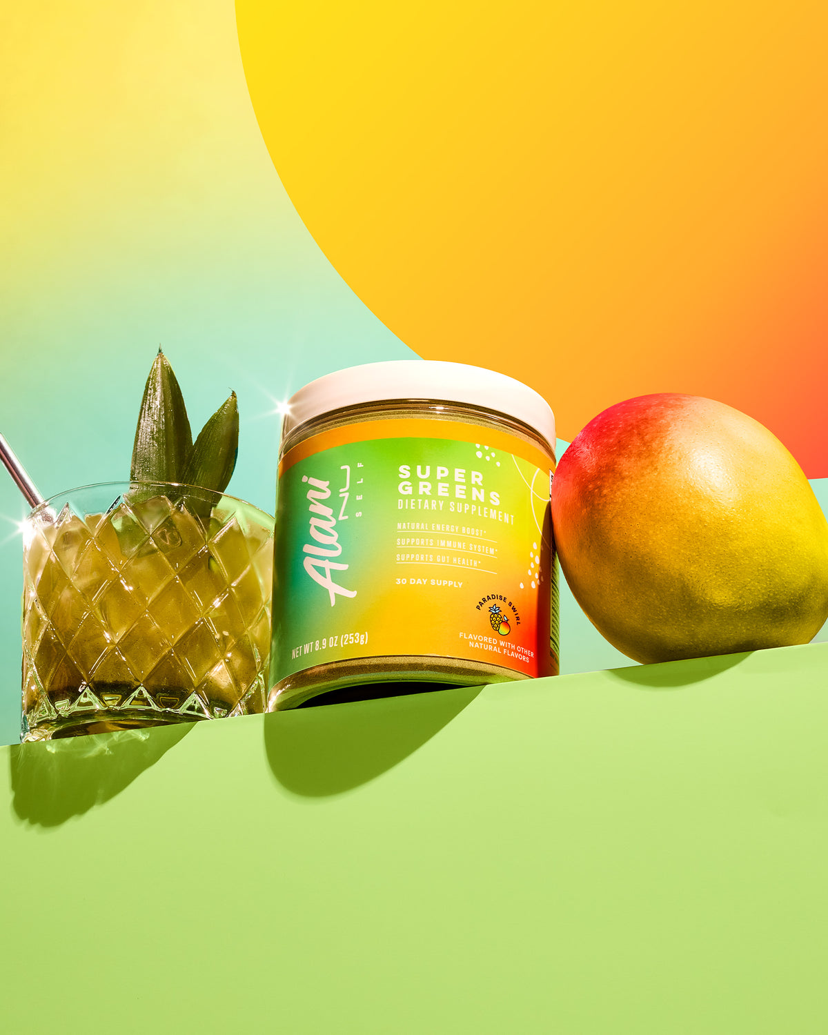 An Alani Nu Super Greens- Paradise Swirl jar sits beside a pineapple-garnished glass and a mango against a vibrant gradient, promising an energy boost while supporting gut and immune health.