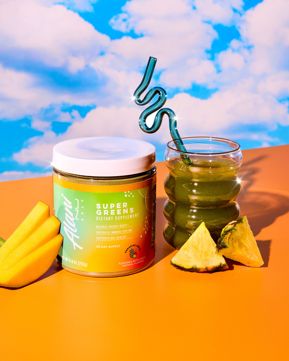 A jar of Alani Nu&#39;s Super Greens- Paradise Swirl supplement sits by a cloudy glass with a wavy straw. Mango slices and pineapple pieces hint at its exotic flavor, offering an energy boost for your gut and immune system.