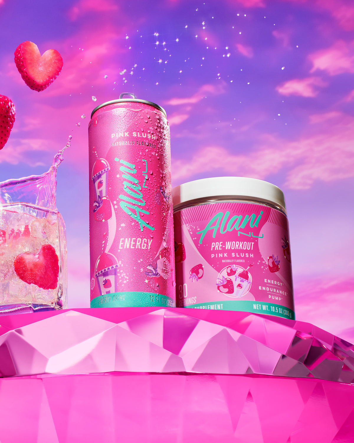 Pink Slush Energy and Pre-Workout side-by-side on a pink-diamond table next to a clear rocks glass with liquid and heart-shaped strawberries splashing upward.  