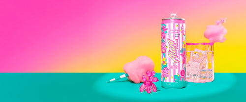 Alani Nu's new Energy Drink poured into a sugar-rimmed glass, spotlighted next to its carnival-inspired can, a cotton candy cone, and a pink balloon dog figurine.


