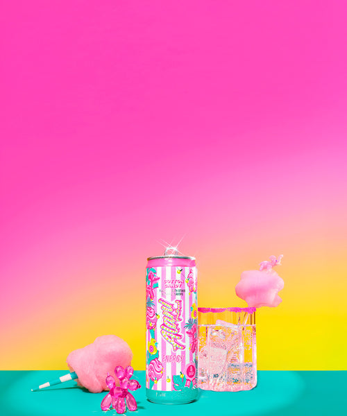 Alani Nu's new Energy Drink poured into a sugar-rimmed glass, spotlighted next to its carnival-inspired can, a cotton candy cone, and a pink balloon dog figurine.
