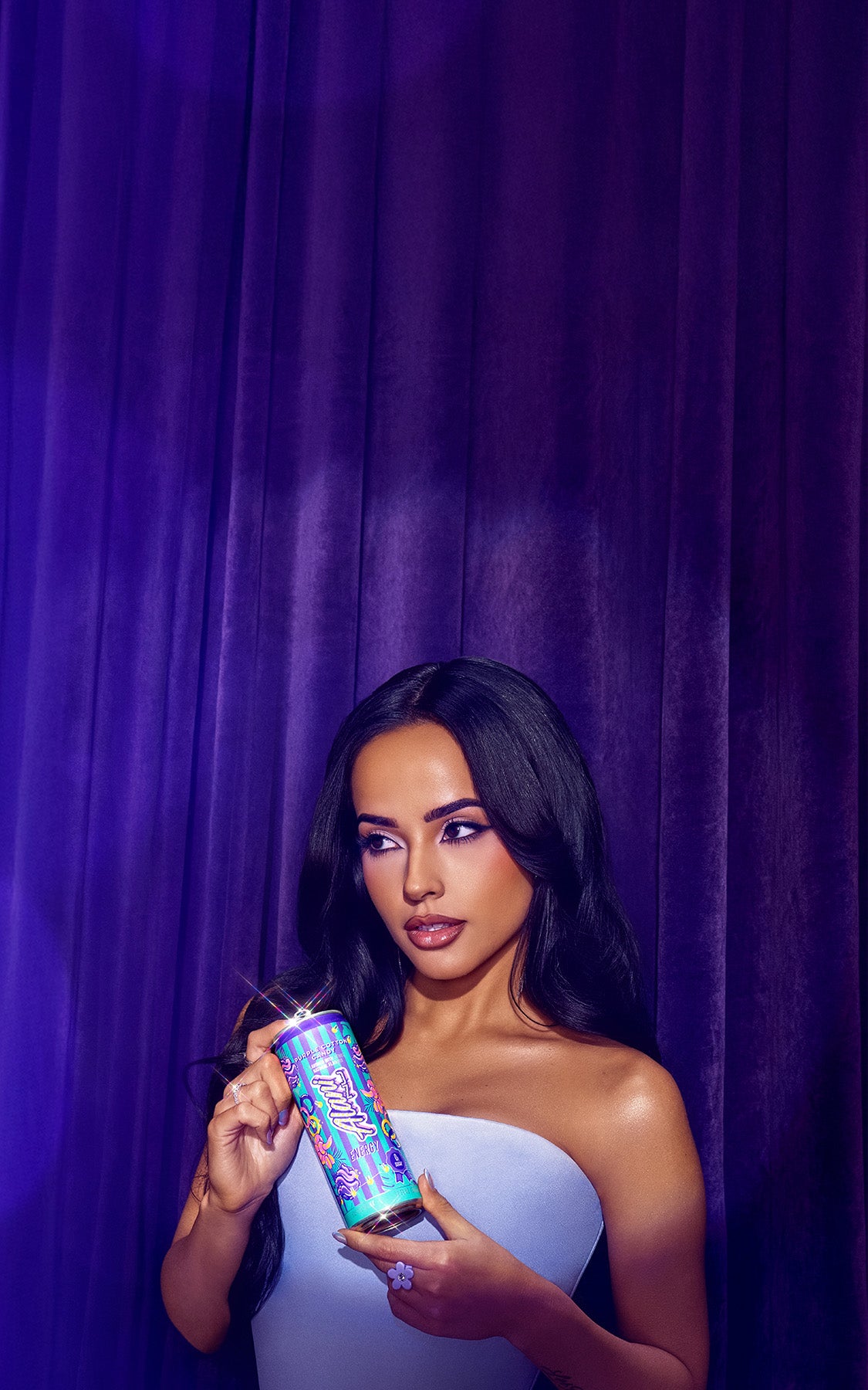 Singer Becky G holding a can of Alani Energy Purple Cotton Candy flavor in front of purple velvet curtains in a soft spotlight.
