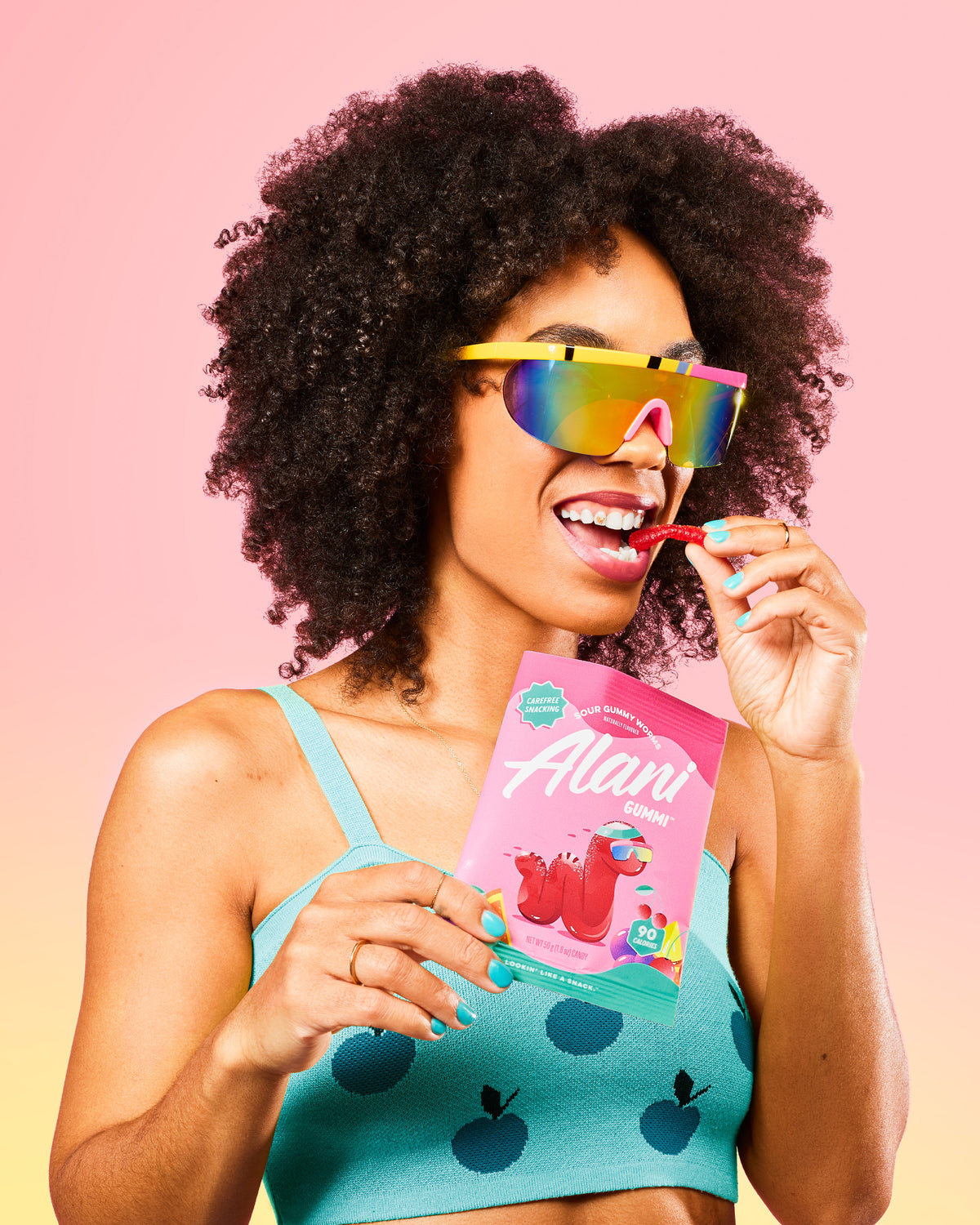 A woman in a bikini top holding a box of Alani Gummi Sour Gummy Worms flavor.