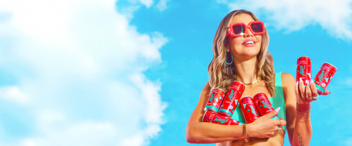 Influencer, Nikki Blackketter, holding an armful of zero-sugar Alani Nu Cherry Slush Energy Drinks—both full-sized cans and minis. 
