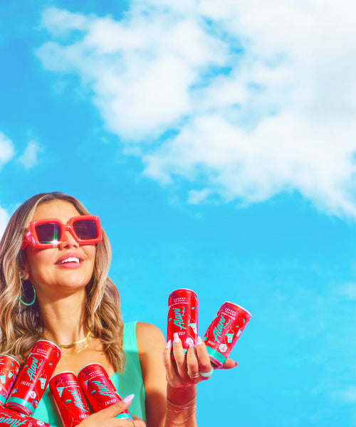 Influencer, Nikki Blackketter, holding an armful of zero-sugar Alani Nu Cherry Slush Energy Drinks—both full-sized cans and minis. 
