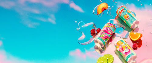 8.4oz Juicy Peach, Cherry Twist, and Sherbet Swirl Mini Energy cans resting on a mirrored surface alongside puddles of Alani Energy and fresh fruit.

