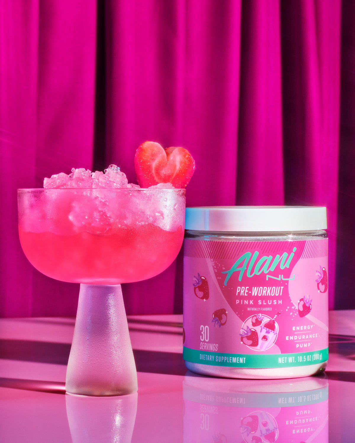 A tub of Alani Nu Pink Slush Pre-Workout next to a frosted coup glass 	filled with pebble ice and pink liquid garnished with a heart-shaped	strawberry.   