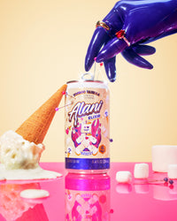 A glossy latex purple hand with a gold and ruby ring reaches down to press a pin into a can of Alani Voodoo Vanilla Energy Elixir, with an overturned ice cream cone with melting soft serve on one side and marshmallows on the other, all reflected on a shiny pink surface against a warm cream background.