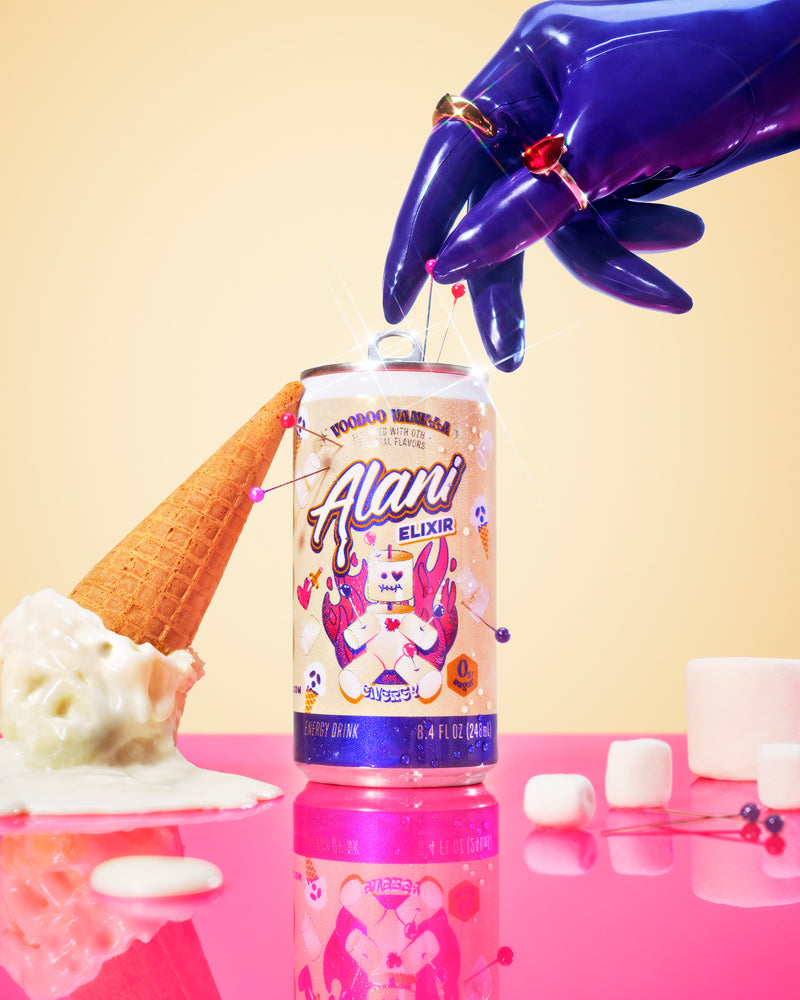 A glossy latex purple hand with a gold and ruby ring reaches down to press a pin into a can of Alani Voodoo Vanilla Energy Elixir, with an overturned ice cream cone with melting soft serve on one side and marshmallows on the other, all reflected on a shiny pink surface against a warm cream background.