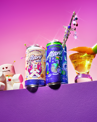 A can of mini Alani Voodoo Vanilla Energy Elixir and a can of mini Alani Witch's Brew Energy balanced on the edge of a purple ledge, next to a marshmallow voodoo doll with pins on one side and a gold cocktail glass garnished with a green apple slice on the other, with a jeweled wand resting inside the mini Witch's Brew can, set against a magenta to pink gradient background.
