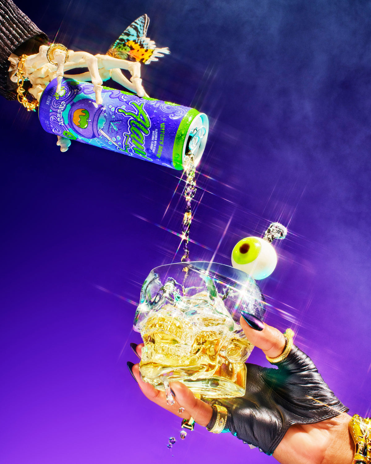 A gold-adorned hand and butterfly-topped figurine pour Alani Nu Energy Drink – Witch’s Brew, with 200mg caffeine and zero sugar, over ice in a gloved hand, topped with a fake eyeball garnish against a purple background.