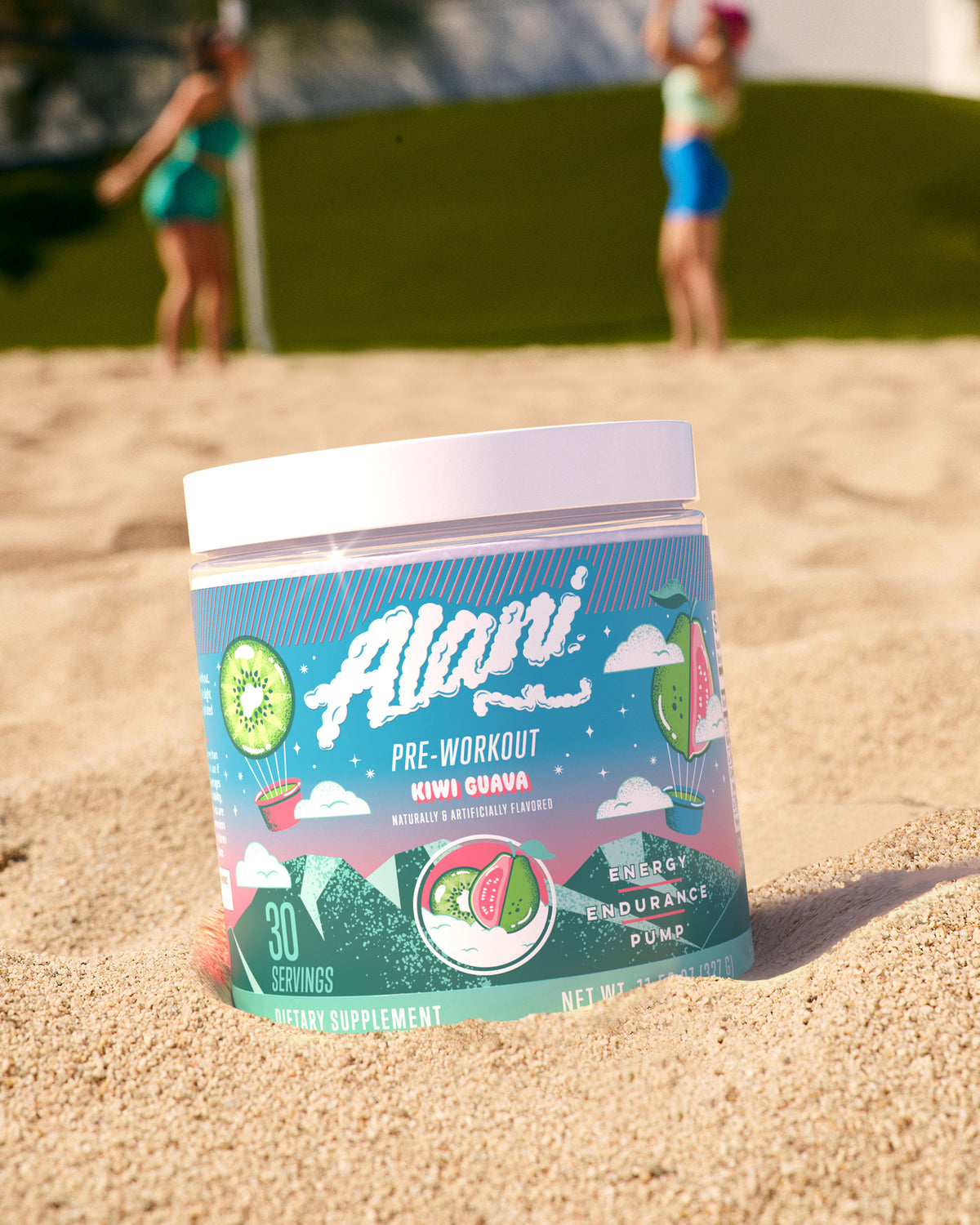 A container of Alani Nu Pre-Workout - Kiwi Guava sits in the sand on a beach.
