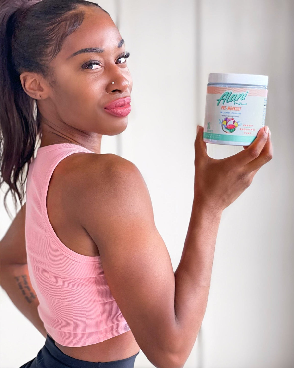 A woman in a pink top holding a container  of Alani Nu Pre-Workout - Island Crush powder.