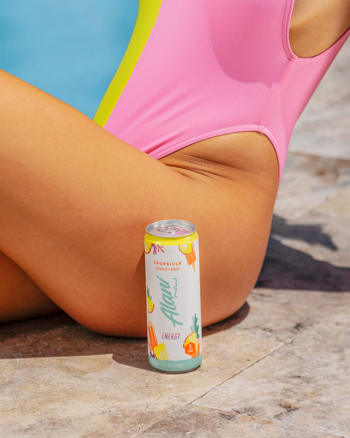 A woman in a pink bathing suit sitting next to a pool with a can of Alani Nu Tropsicle Energy Drink.