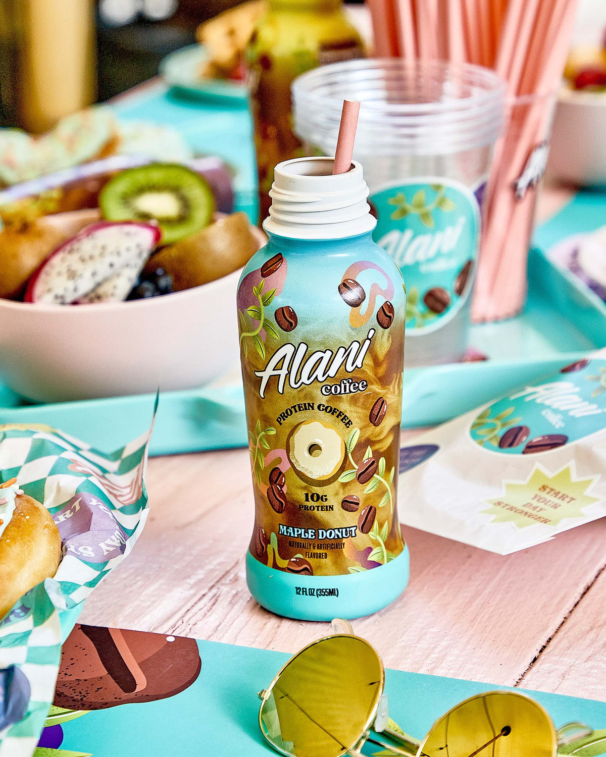 A bottle of Alani Nu Coffee - Maple Donut is on a table with a bowl of fruit in the background.